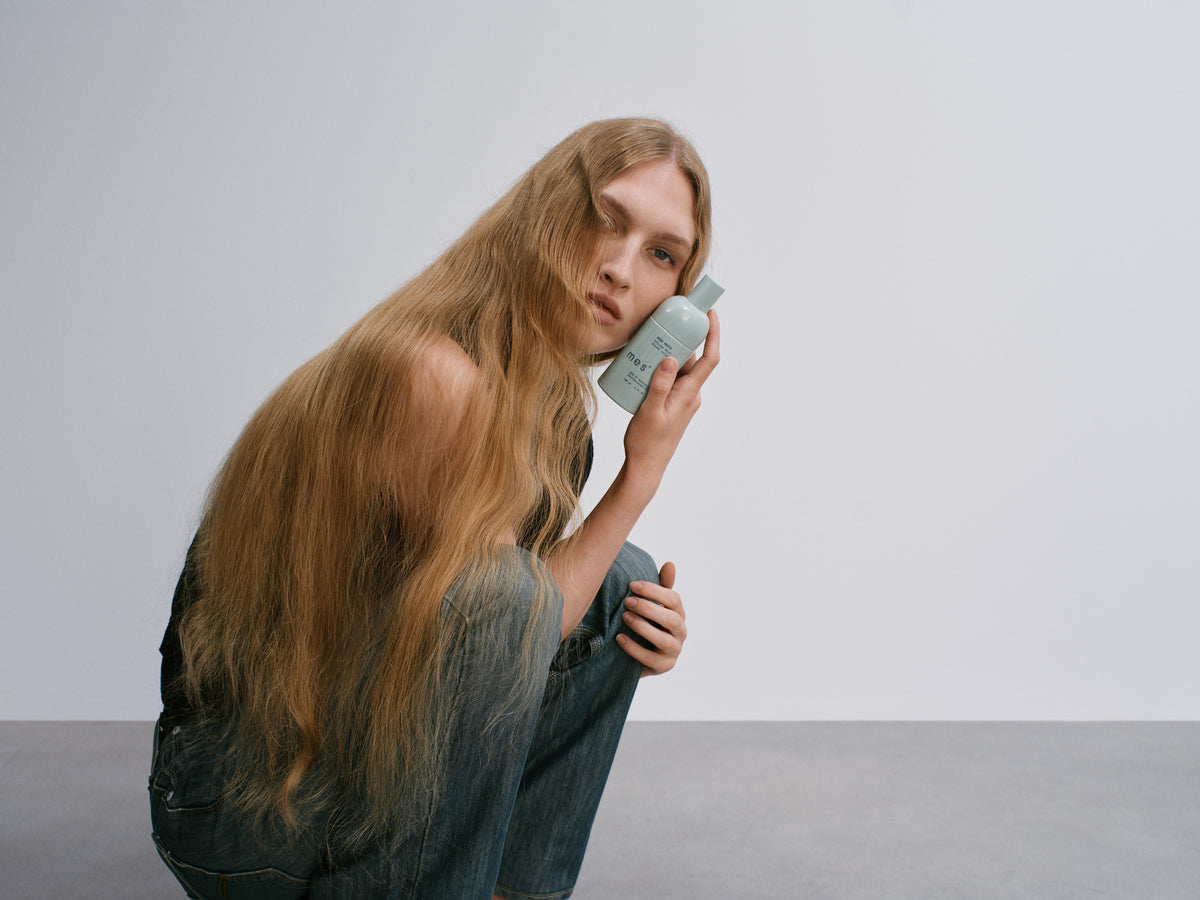 A woman with long blonde hair crouching, holding Good Roots Volume Mist.