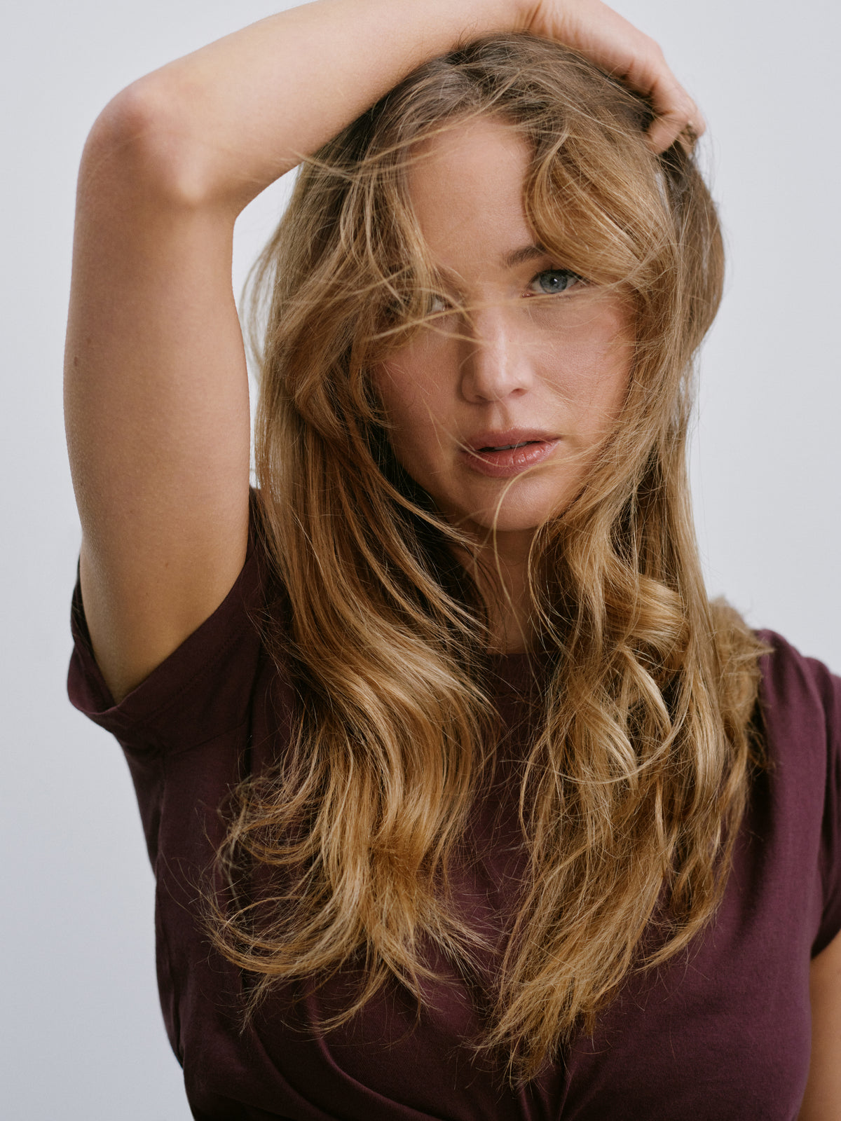 Jennifer Lawrence in a burgundy top, one hand in her long wavy hair.
