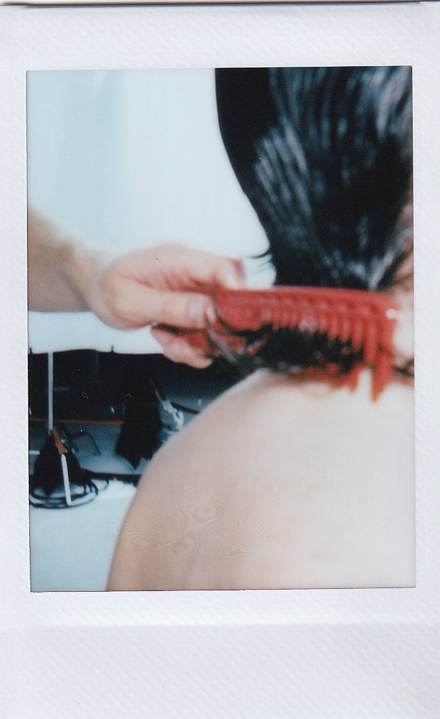 Polaroid of a hand combing through long dark hair with a red comb.